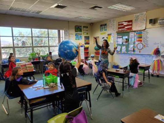 A female teacher with long curly brown hair and light skin passes a beach ball of the Earth to students seated in a classroom